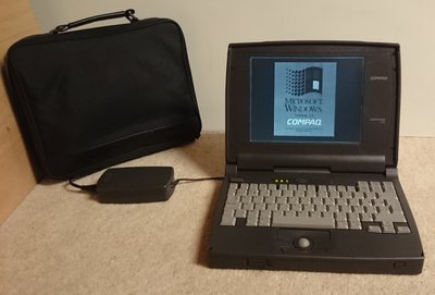 Compaq Contura 400 ouvert affichant l'écran de démarrage Windows 3.1, sacoche de transport et bloc d'alimentation posés à côté
