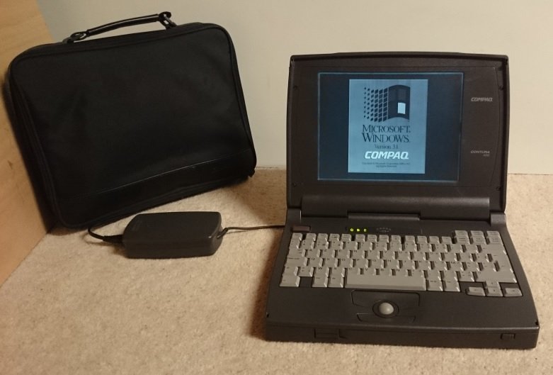 Compaq Contura 400 ouvert affichant l'écran de démarrage Windows 3.1, sacoche de transport et bloc d'alimentation posés à côté