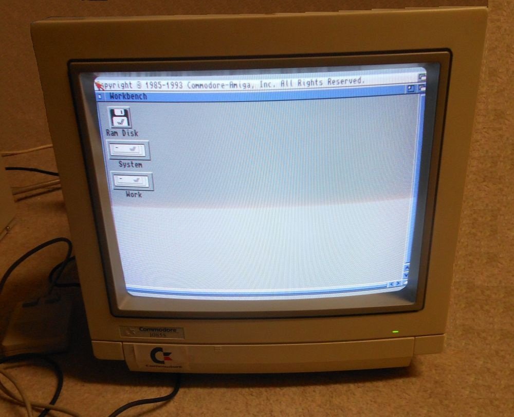 Moniteur Commodore relié à un Amiga 2000, affichant le bureau Workbench avec les icônes Ram Disk, System et Work