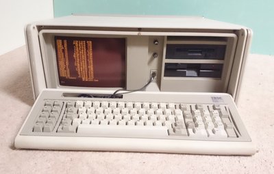 IBM 5155 ouvert, écran ambre intégré affichant du texte, clavier IBM posé devant l'unité centrale