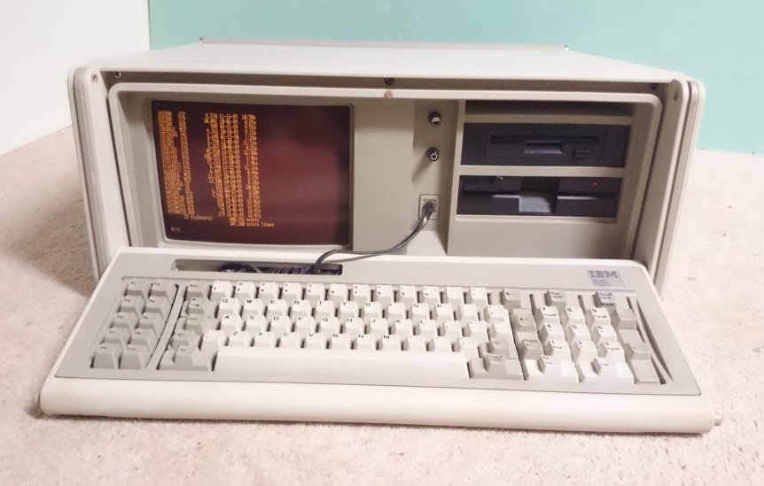 IBM 5155 ouvert, écran ambre intégré affichant du texte, clavier IBM posé devant l'unité centrale