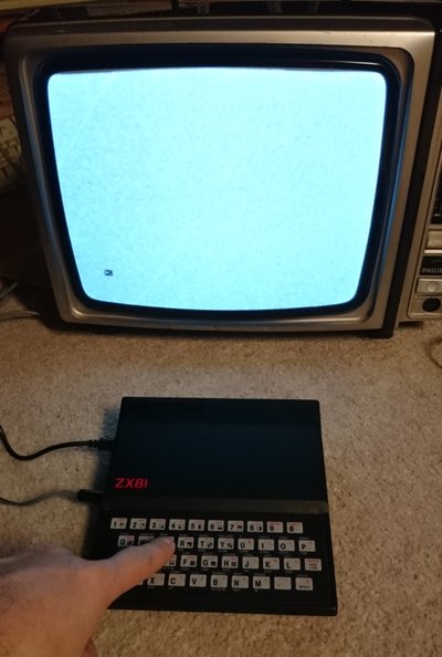 Sinclair ZX81 posé devant un téléviseur à tube affichant un signal test, une main appuyant sur une touche du clavier
